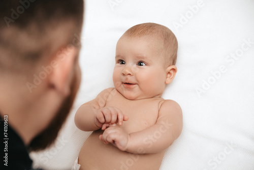 The baby is lying in a diaper on a white blanket, looking at dad and smiling. Top view. 