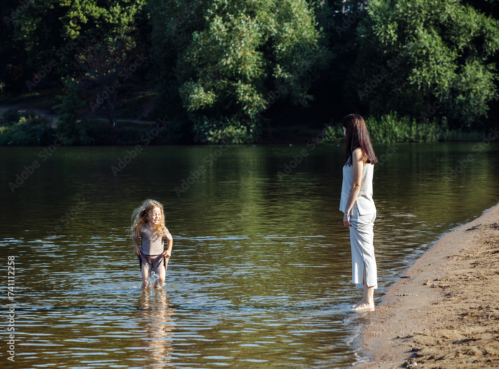 Fototapeta premium A five-year-old cheerful girl and her mother are walking by the lake, barefoot in the water, having fun on a summer day