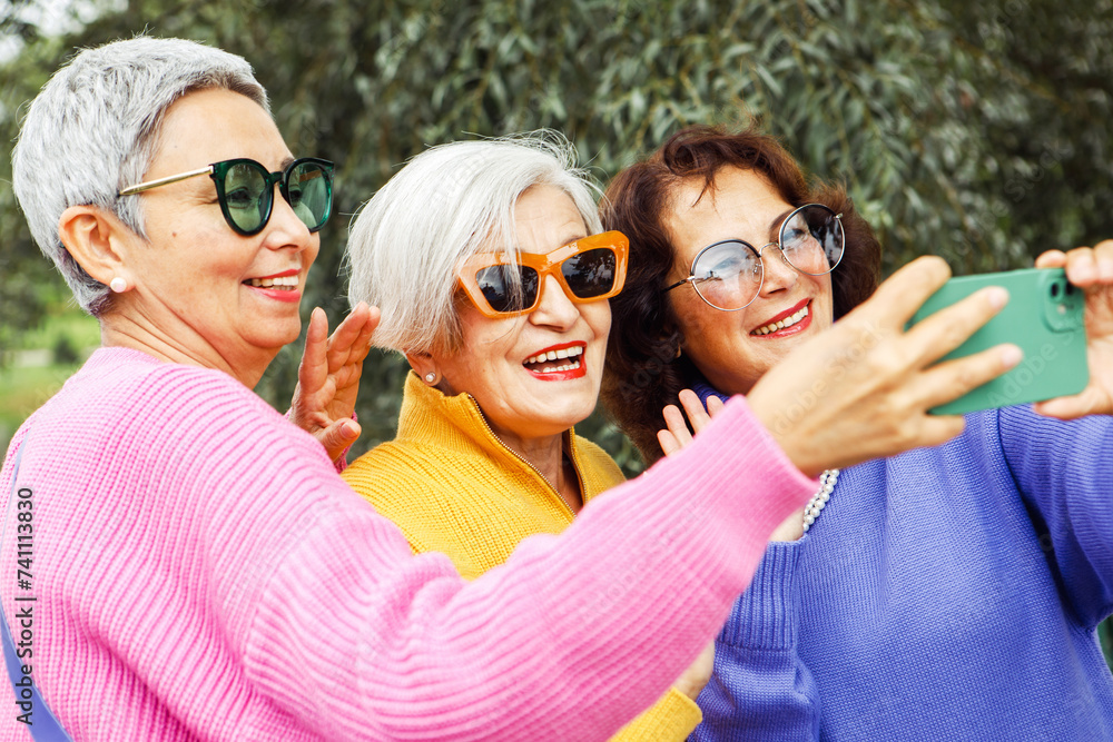 Three cheerful elderly friends in bright sweaters take a selfie on a smartphone and laugh, having a great time. Three cheerful elderly friends in bright sweaters take a selfie on a smartphone and laugh, having a great time.