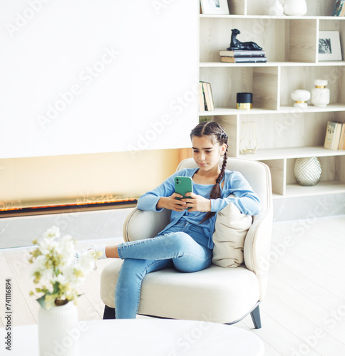 Free Download Little kid cute girl sitting on chair, having fun at home, using smartphone, playing video game on mobile phone. - GFXLoad