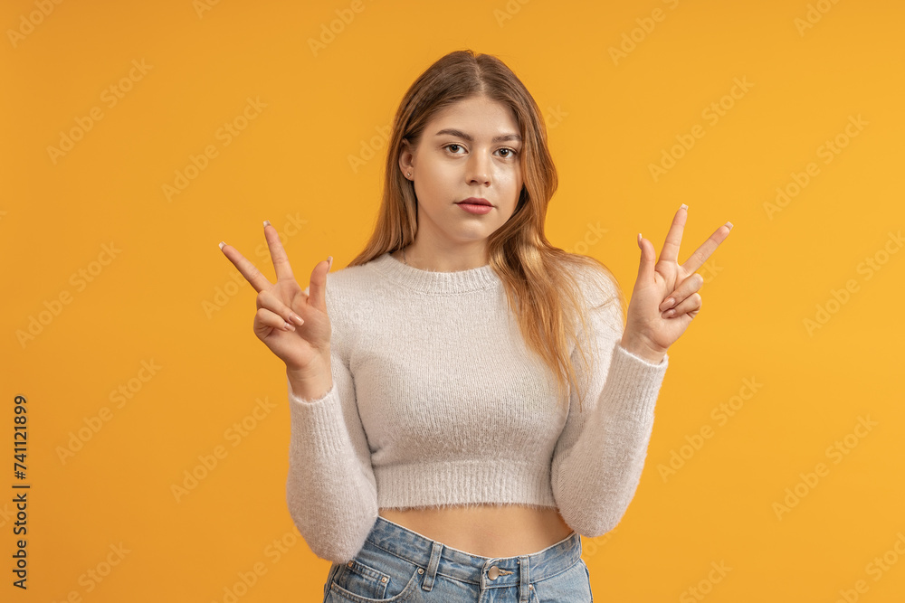 A young woman in a white cropped sweater on a yellow background holding up peace signs with both hands. A young woman in a white cropped sweater on a yellow background holding up peace signs with both hands.