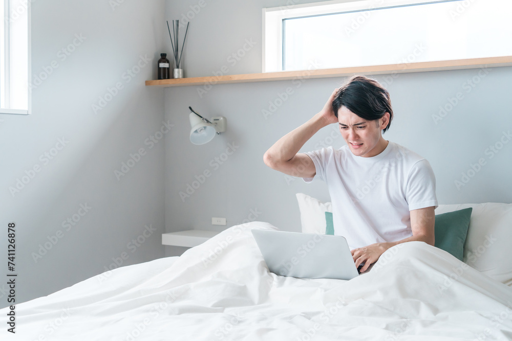 家・ホテルの寝室・ベッドルームでノートパソコンを使う若い男性（困る・悩む・ストレス・トラブル）
