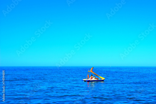 Delightful scene from Portonovo in Riviera del Conero with the intensely rippling blue waters of the Adriatic Sea surrounding a lone white and blue pedalo with a yellow slide, children and women