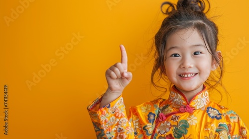 5 year old girl in a fresh and neat cheongsam Smiling brightly as he pointed up and to the right. With a bright yellow backdrop. It adds a delightful touch.