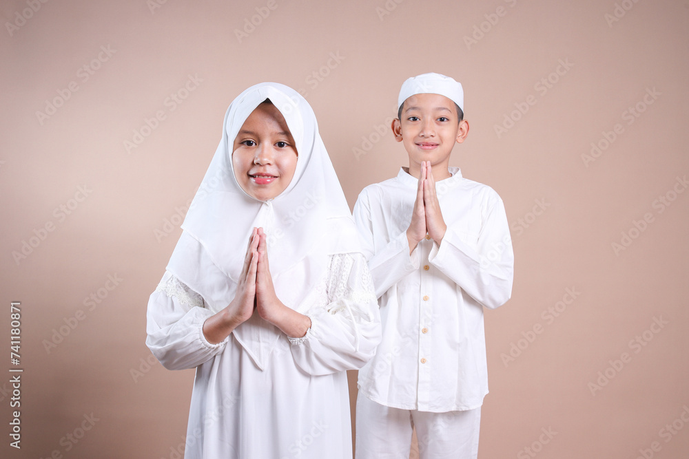 Asian muslim boy and sister clasped their hands showing greeting gesture over beige background Asian muslim boy and sister clasped their hands showing greeting gesture over beige background