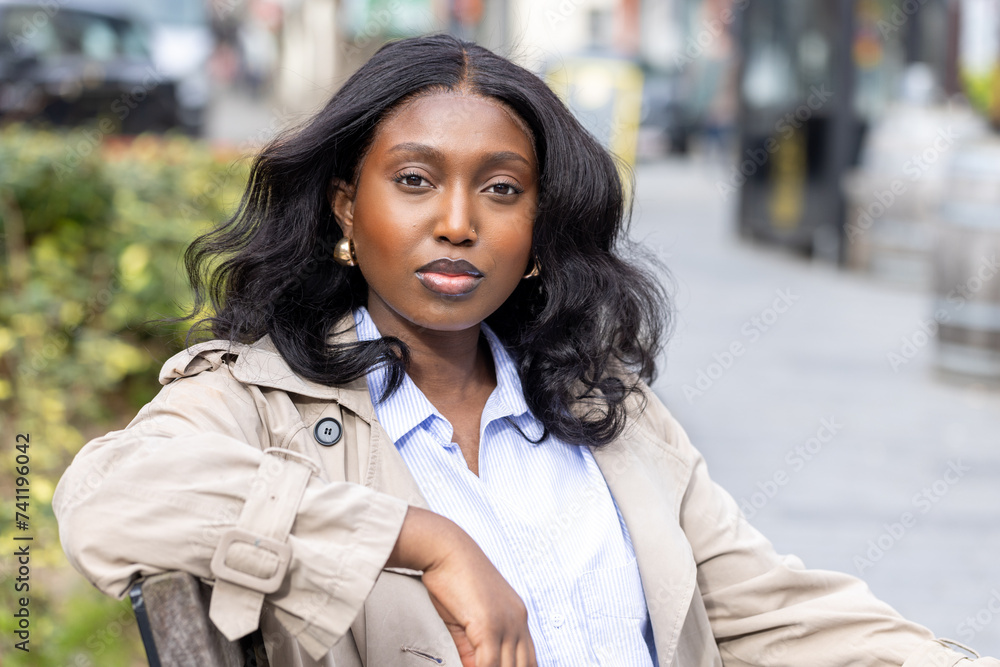 This portrait features a young African woman seated confidently in an urban setting. She is dressed in smart casual attire, with a light trench coat over a blue-striped shirt, perfect for a This portrait features a young African woman seated confidently in an urban setting. She is dressed in smart casual attire, with a light trench coat over a blue-striped shirt, perfect for a