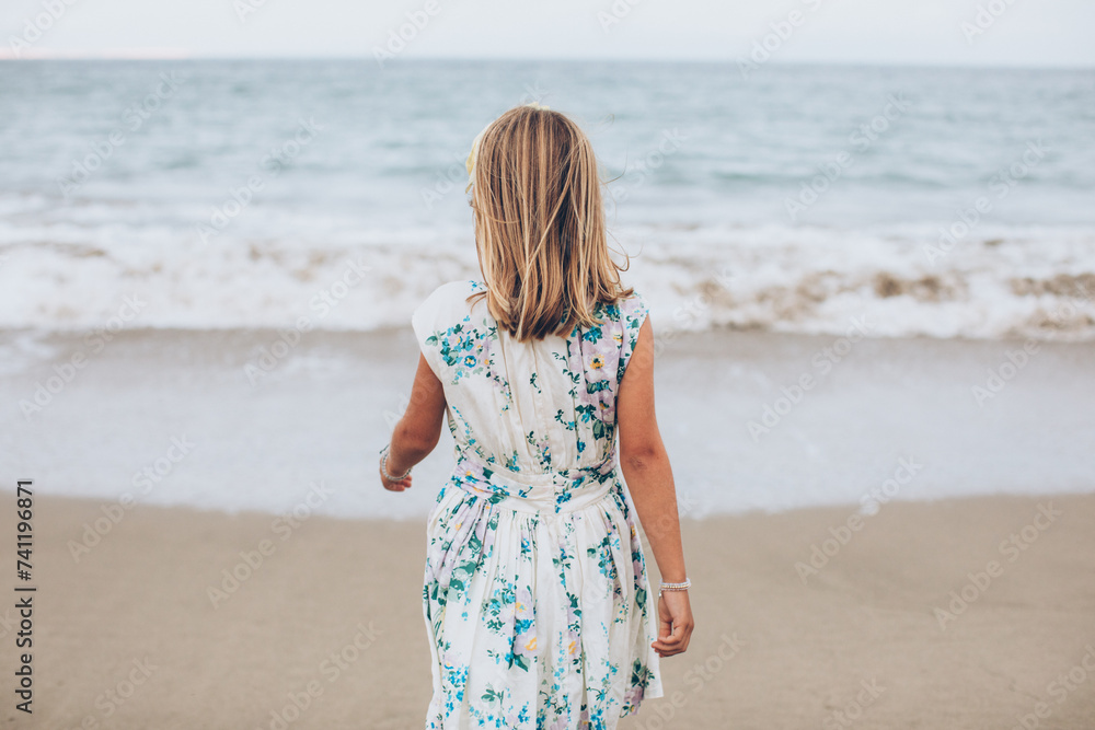 young girl looking out at the ocean walking on the coastline and wearing a blue floral dress young girl looking out at the ocean walking on the coastline and wearing a blue floral dress