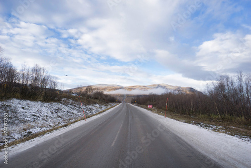 An open road stretches toward a snow-capped mountain under a partly cloudy sky, evoking a sense of travel.