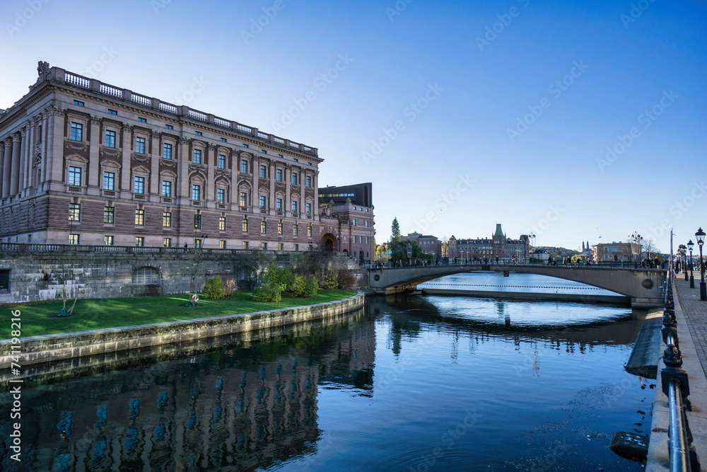 A serene riverscape with historical buildings reflected in the water, under a blue sky in a tranquil city setting.