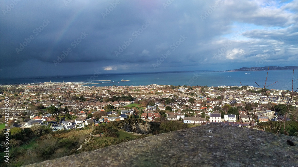 Panoramic view of a town by the sea under dramatic skies with a visible rain shaft and a rainbow.