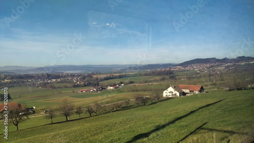 Countryside view from a train window showing farmland, houses, and distant hills under a clear blue sky.