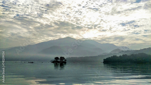 Fototapeta Naklejka Na Ścianę i Meble -  A misty landscape with a mountain silhouette, a lone island with trees, and a textured sky reflecting on calm water.