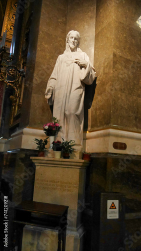 A white marble statue of a religious figure with a heart and flowers placed at its base, inside a church.