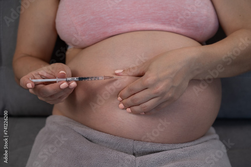 A pregnant woman puts an injection of insulin while sitting on the couch. Close up of the belly.