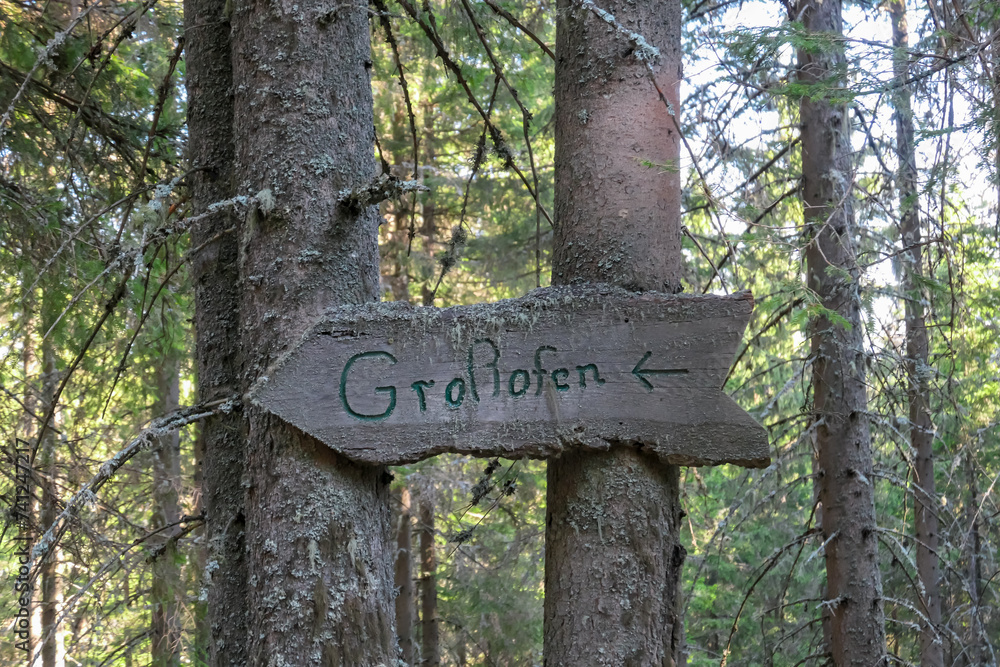 Directional path sign leading to unique rock formations Grossofen surrounded by idyllic forest in Modriach, Hebalm, Kor Alps, border Carinthia Styria, Austria. Wooden board fixed on large tree