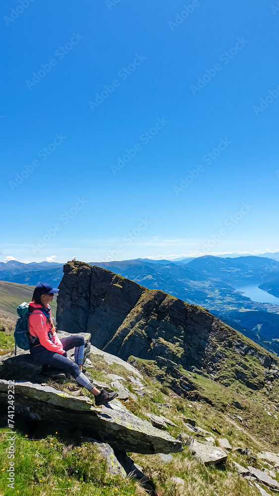 Hiker woman sitting on rock on alpine meadow with panoramic view of lake Millstatt. Idyllic hiking trail to mountain peak Boese Nase, Ankogel Group, Carinthia, Austria. Remote Austrian Alps in summer