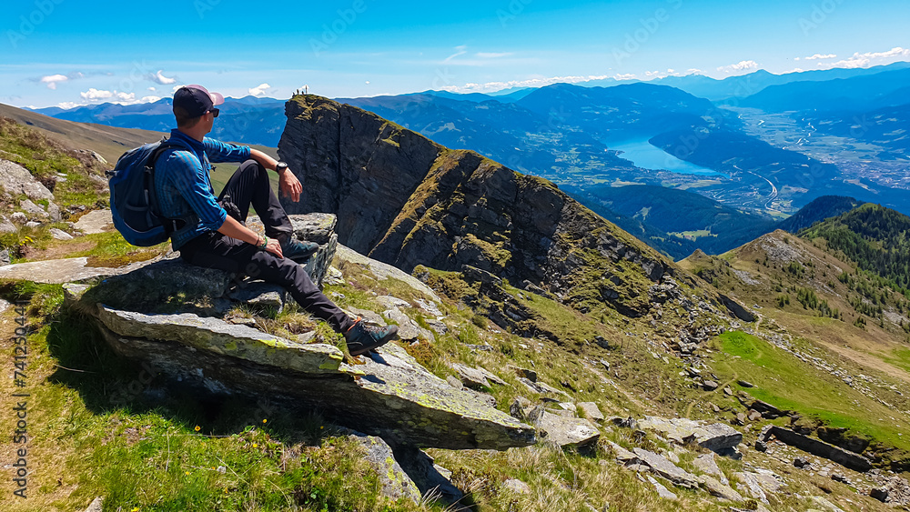 Hiker man sitting on rock on alpine meadow with panoramic view of lake Millstatt. Idyllic hiking trail to mountain peak Boese Nase, Ankogel Group, Carinthia, Austria. Remote Austrian Alps in summer Hiker man sitting on rock on alpine meadow with panoramic view of lake Millstatt. Idyllic hiking trail to mountain peak Boese Nase, Ankogel Group, Carinthia, Austria. Remote Austrian Alps in summer