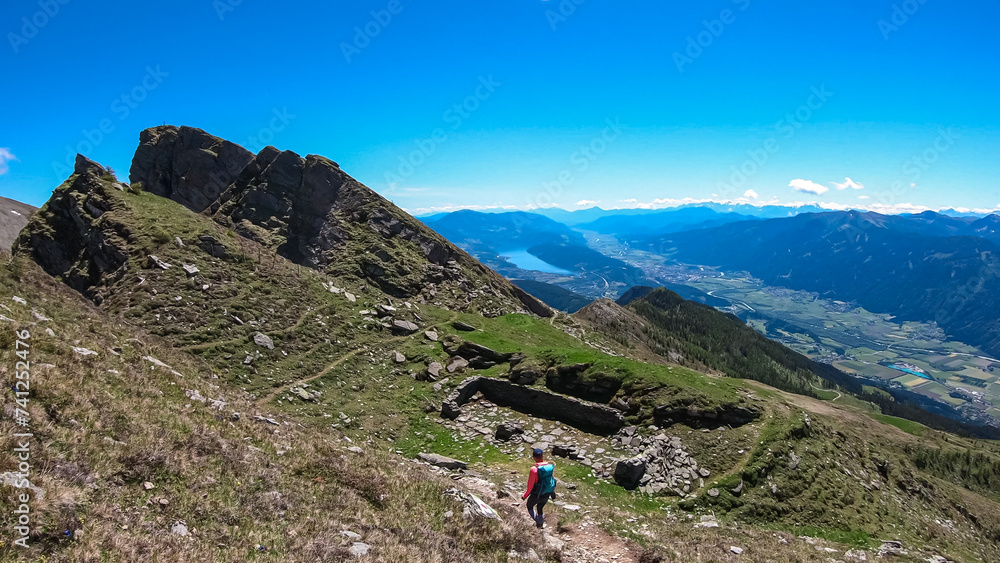 Hiker woman on idyllic hiking trail on alpine meadow with scenic view of lake Millstatt seen from mountain peak Boese Nase, Ankogel Group, Carinthia, Austria. Remote landscape. Austrian Alps in summer