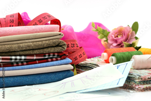 Stack of different fabrics and accessories for sewing on a white background. Lots of fabric, spools of thread, measuring tape and patterns for sewing clothes. Sewing process.