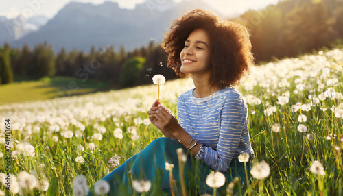 Beautiful Young Woman sitting on the field in green grass and blowing dandelion. Outdoors. Enjoy Nature. Healthy Smiling Girl on summer lawn. Allergy free concept. Gorgeous slim mixed race Caucasian