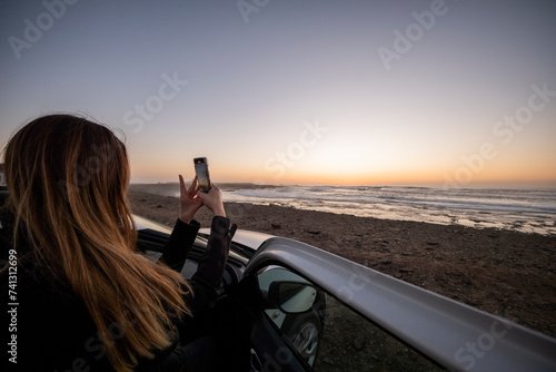 Chica desde su coche de alquiler de vacaciones sacando una foto de la puesta del sol en la playa de Fuerteventura photo