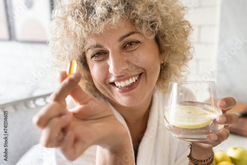 Woman Healthcare. Female Holding Suplement in a Bright Room. Portrait of a joyful curly-haired woman taking a vitamin pill with a glass of water. Concept of health, wellness, and daily routine 
