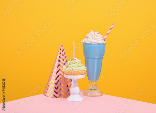 Sweet desserts and a holiday hat. Blue milkshake with bright sprinkles and straw, cupcake with whipped cream and birthday candle.