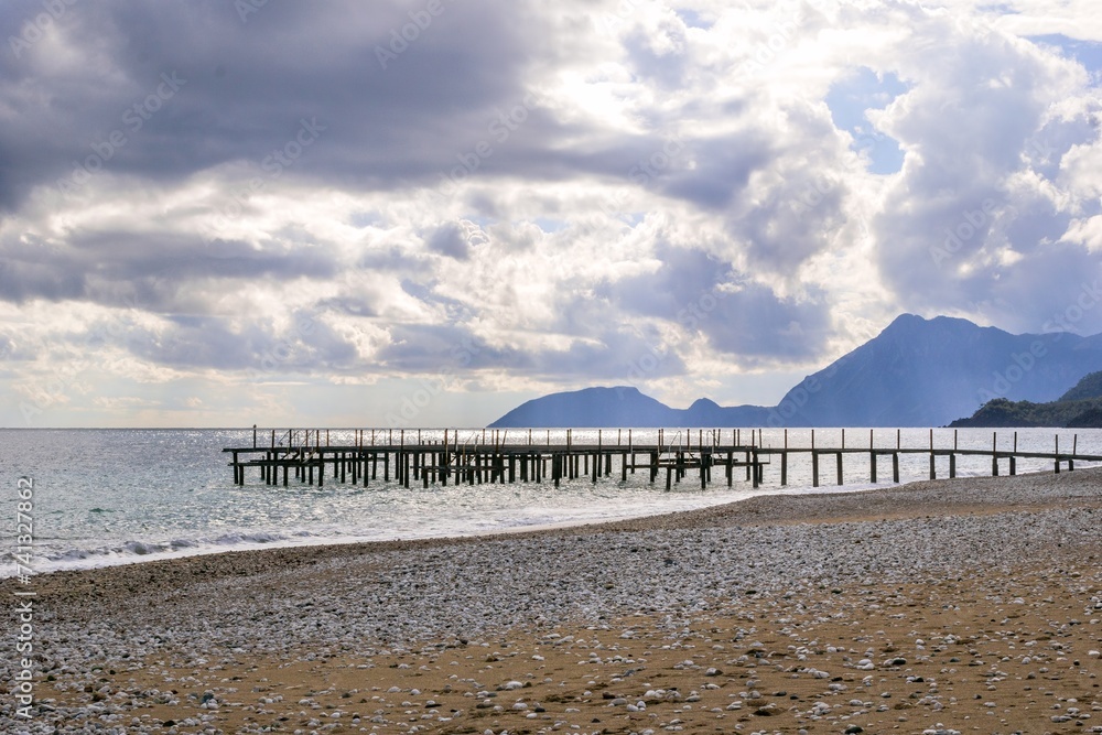 Tranquil Wooden Dock Extending into Calm Blue Sea with Mountain Range under Cloudy Sky