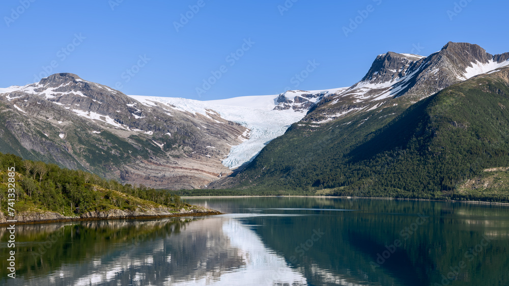 A panoramic display of the Svartisen Glacier descending into lush greenery, with its beauty mirrored in the glassy fjord, encapsulates the serenity of the Arctic