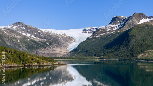 A panoramic display of the Svartisen Glacier descending into lush greenery  with its beauty mirrored in the glassy fjord  encapsulates the serenity of the Arctic