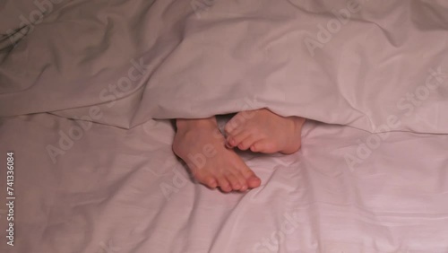 kid boy legs itching and scabies in bed in the bedroom in the evening. Restless baby child girl scratches her feet with fungus on the sheet under the blanket