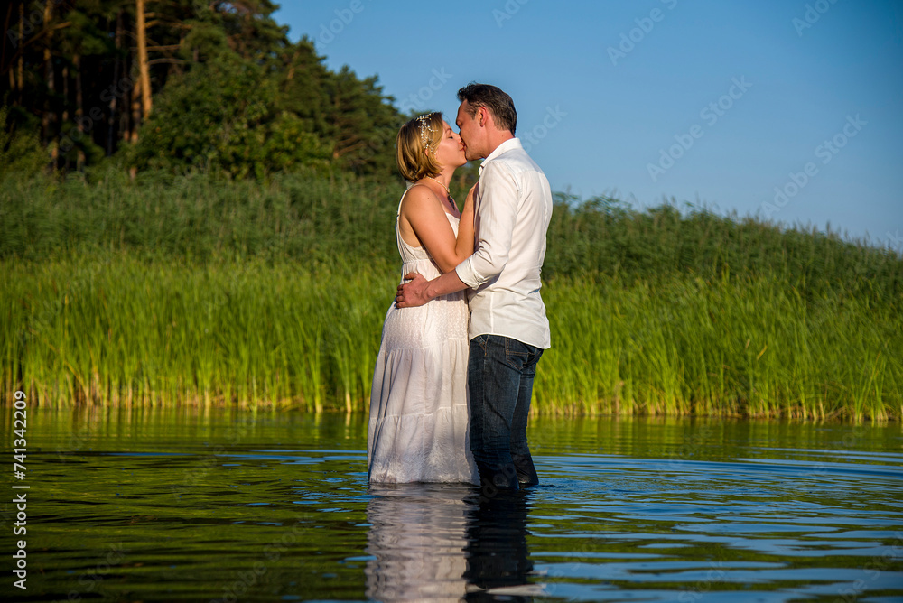 beautiful  happy hugging couple, bride and groom in white dresses walking in forest river on sunny summer day and holding each other hand by hands