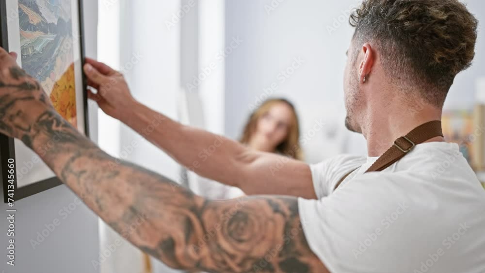 A tattooed man and spectacled woman collaborate in hanging a framed painting in a bright art studio. A tattooed man and spectacled woman collaborate in hanging a framed painting in a bright art studio.