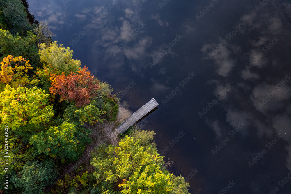 A drone flies over a huge blue lake with a wooden pier among the forests on a cloudless autumn day at dawn.
