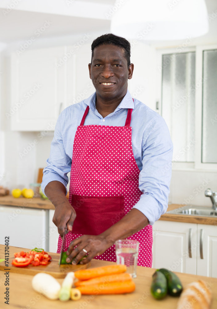 Smiling adult man in red apron preparing vegetable dish in home kitchen
