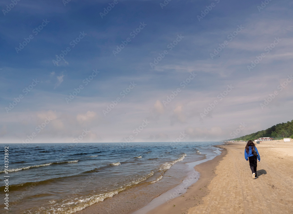 Teenager girl walking on yellow sandy beach of Jurmala, Latvia, popular tourist landmark area with great vibe and clean air. Cloudy sky. Coast line with blue water and sky. Teenager girl walking on yellow sandy beach of Jurmala, Latvia, popular tourist landmark area with great vibe and clean air. Cloudy sky. Coast line with blue water and sky.