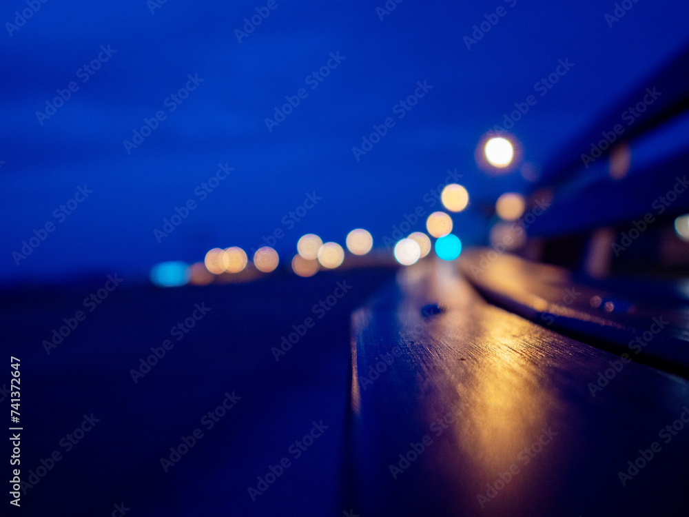 City lights illuminate wooden bench. Blue night sky with town lights out of focus in the background. Selective focus. Abstract night city life.