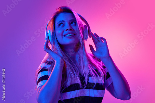 Gel portrait of beautiful young woman listening to music in headphones with dreamy expression against pink studio background in neon light. Concept of human emotions, youth, fashion, expression