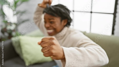 A young black woman with dreadlocks excitedly crosses her fingers while sitting on a sofa indoors, showcasing anticipation or hope. photo