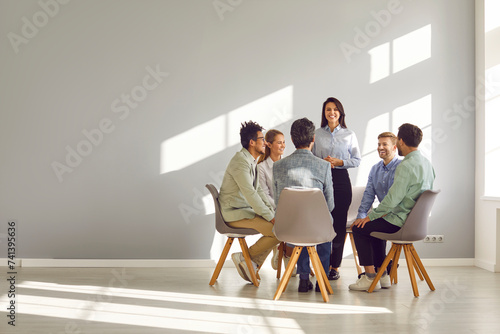 Corporate business team meeting with a manager or coach. Group of happy diverse, multiracial people sitting in a circle around a standing woman in an office with a light grey copy space wall © Studio Romantic