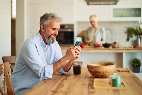 A cheerful senior adult man waiting for breakfast at a dining table and using his phone