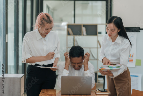 Stressed asian businessman with hands on head in a meeting as female colleagues discuss documents. Workplace stress and deadline pressure concept.