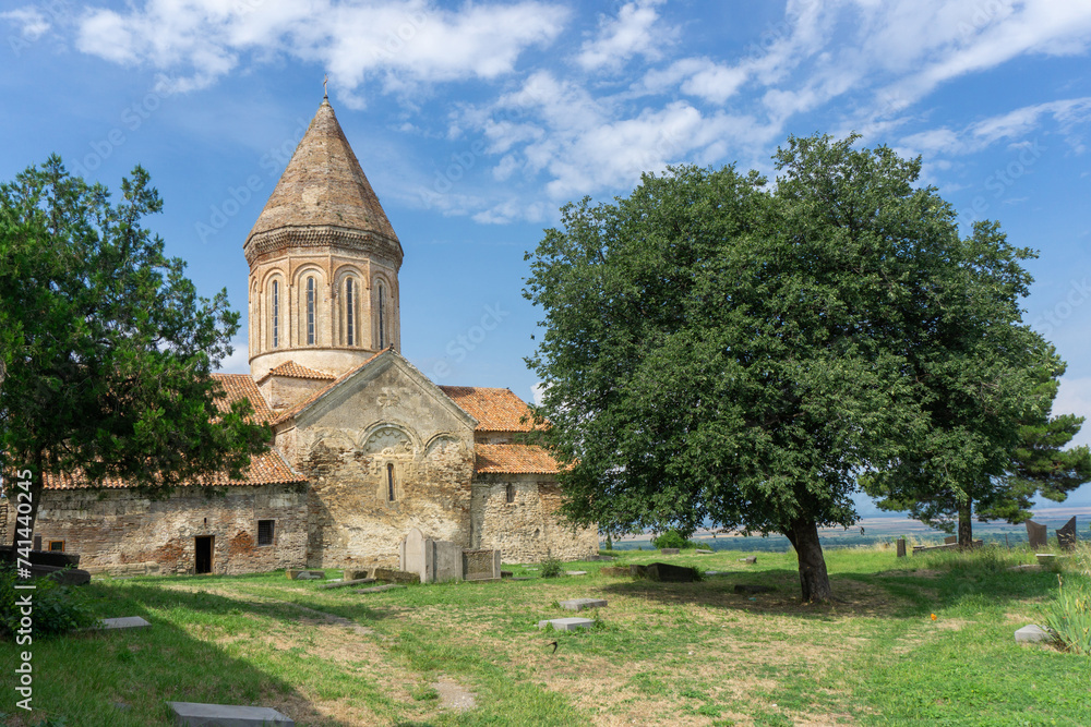 Church of Khirsa monastery of Saint Stephen. Stone walls, orange tile roof, green lawn, blue bright sky with clouds, gravestones and huge tree. Visible Alazani valley Church of Khirsa monastery of Saint Stephen. Stone walls, orange tile roof, green lawn, blue bright sky with clouds, gravestones and huge tree. Visible Alazani valley
