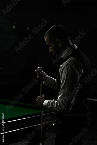 Fotografie Dark image of young man in classical wear preparing cue with chalk before snooker game
