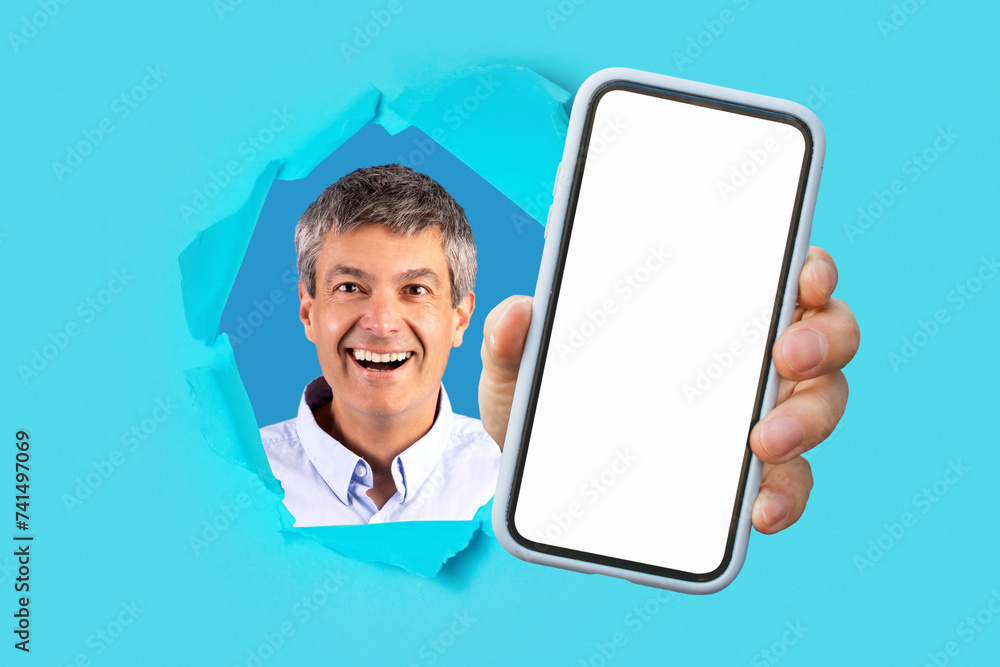 Mature man with a friendly smile peeking through a torn blue paper, presenting a smartphone Mature man with a friendly smile peeking through a torn blue paper, presenting a smartphone