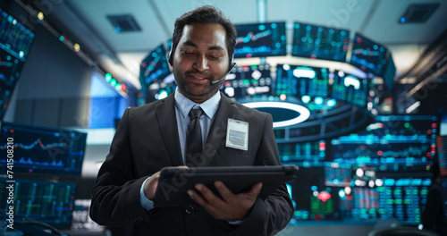 Portrait of a Young Indian Man Working in an International Stock Exchange Company: Happy Trader Using a Tablet Computer, Monitoring, Talking to a Corporate Business Partner in Headphones