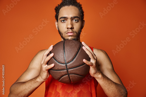 young sporty african american man holding basketball and looking at camera on orange backdrop © LIGHTFIELD STUDIOS