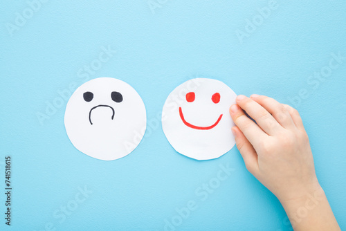 Baby hand with bad and happy smileys on light blue table background. Pastel color. Negative and positive smiling face expressions on white paper. Closeup. Children mood concept. Top down view.
