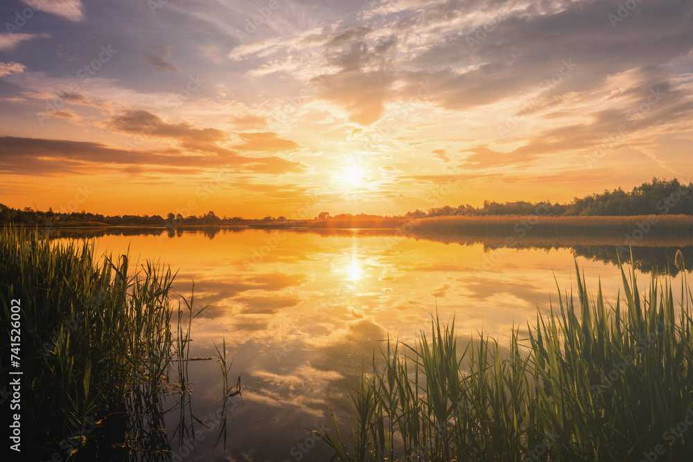Sunset or sunrise above the pond or lake at spring or early summer evening or morning with cloudy sky and reed grass. Springtime landscape. Water reflection. Vintage film aesthetic. - obrazy, fototapety, plakaty 