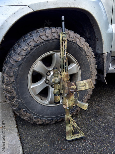 A camouflage-colored rifle gun standing against a mud-covered wheel of a silver SUV